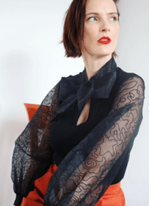 Black top featuring sheer, voluminous sleeves with intricate abstract patterns and a coordinating necktie, paired with high-waisted rust-colored pants for a sophisticated, modern look.