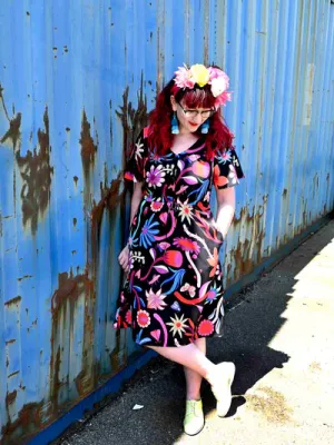 Woman dressed in a black dress with large, bold floral designs in reds, blues, and oranges, styled with a flower crown and leaning casually against a weathered blue wall. Designed by Australian designer brand Streborclothing.