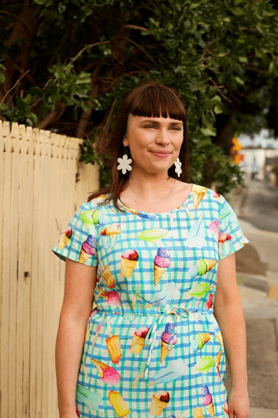 Woman wearing a green and blue dress with a colorful abstract print featuring fruit and ice-cream illustrations, styled for a playful, sunny-day look.