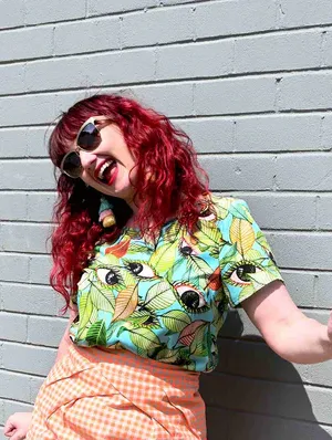 A short-sleeved top with a bold leaf and seed pattern in vibrant green, orange, and black on a light blue background, displayed against a pink backdrop to highlight its colorful appeal. Designed by Australian designer brand Streborclothing.