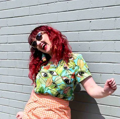 A short-sleeved top with a bold leaf and seed pattern in vibrant green, orange, and black on a light blue background, displayed against a pink backdrop to highlight its colorful appeal. Designed by Australian designer brand Streborclothing.