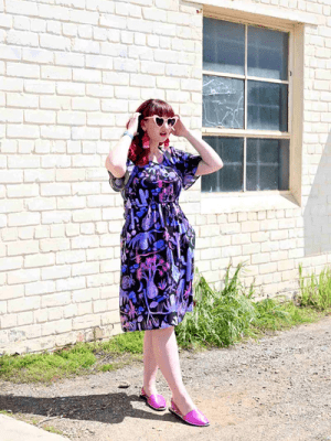 Woman wearing a dark dress with a whimsical cactus and desert-themed print in pinks, purples, and blues, standing against a graphic backdrop with matching patterns. Designed by Australian designer brand Streborclothing.