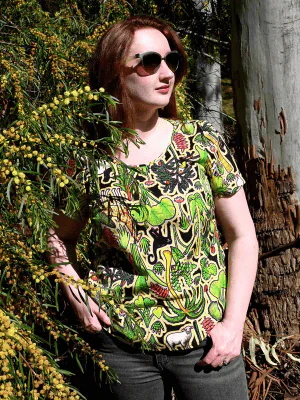 This top showcases a complex pattern with elements of flora, including leaves, flowers, and abstract natural shapes in shades of green, red, and yellow. The design appears dense and rich, resembling an intricate forest scene. Designed by Australian designer brand Streborclothing.