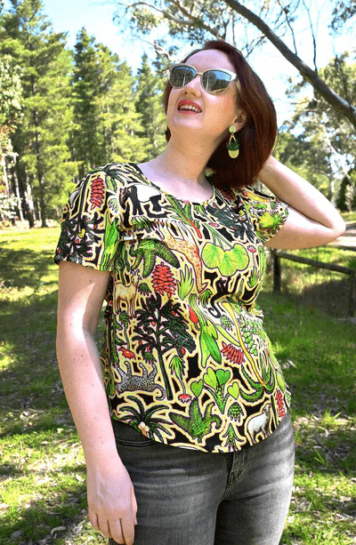 This top showcases a complex pattern with elements of flora, including leaves, flowers, and abstract natural shapes in shades of green, red, and yellow. The design appears dense and rich, resembling an intricate forest scene. Designed by Australian designer brand Streborclothing.